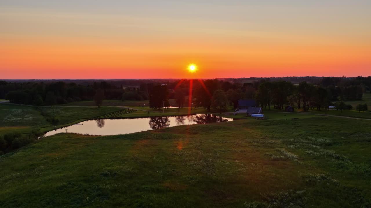 Latvian countryside village house by the pond. Aerial slow flying forward towards sun setting over a tranquil pond and rural landscape in Latvia, with warm orange skies reflecting in the water -