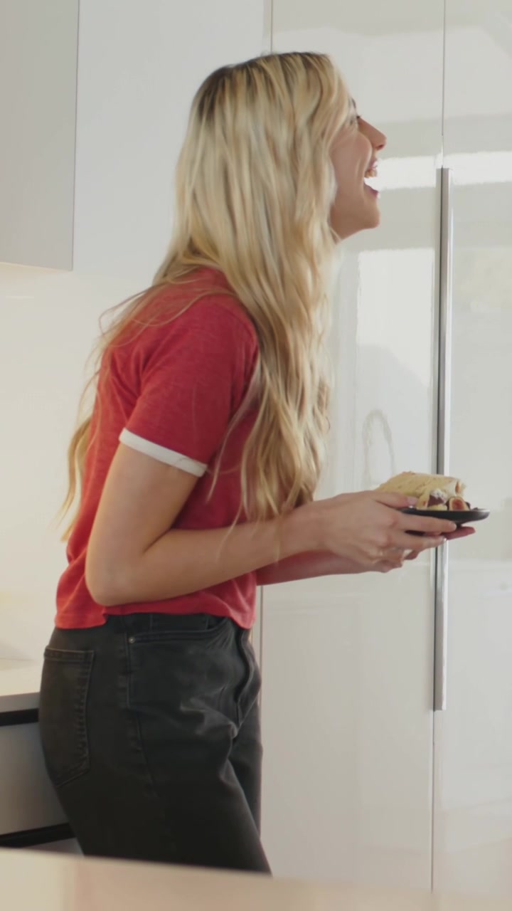 Vertical video: Laughing after remark, woman in red top leaning at home counter eating from plate