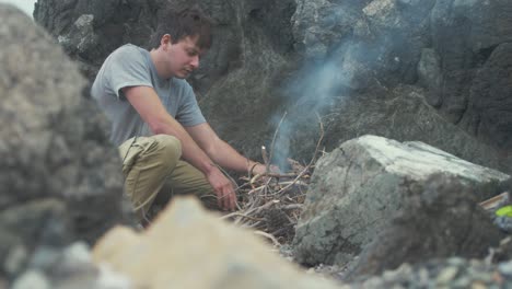 young man lighting campfire alone on rocky seaside shoreline