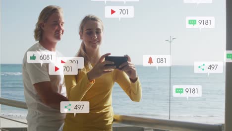 Animation-of-social-media-notifications-over-smiling-couple-taking-selfie-on-holiday-by-the-sea