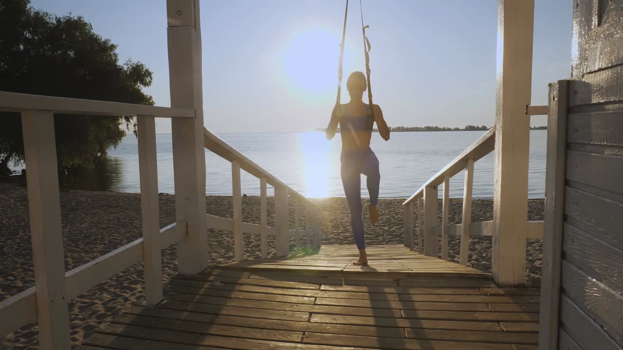 야외 운동. 서스펜션 스트. 스트으로 운동. 운동하는 젊은 여성은 줄을 사용하여 전신 저항 운동을 하고 있습니다. 해가 지거나 해가 뜨는 동안 해변에서. 건강한 생활 방식 개념. 아침 스포츠.