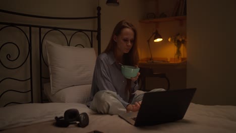 woman eating breakfast in bed while working on laptop at night