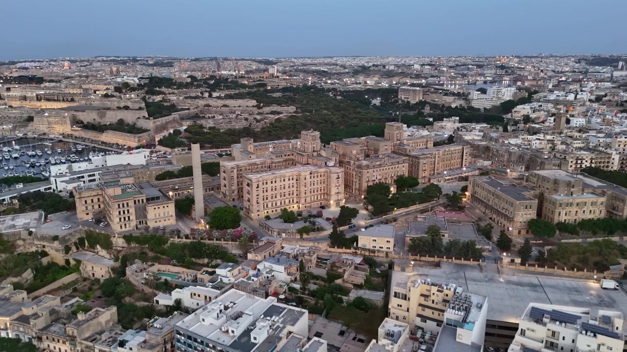 Skyline di Pietà, Malta, con l'Ospedale San Luca, il porto turistico, edifici residenziali e storici, strade e vegetazione lungo la costa mediterranea in una veduta aerea serale