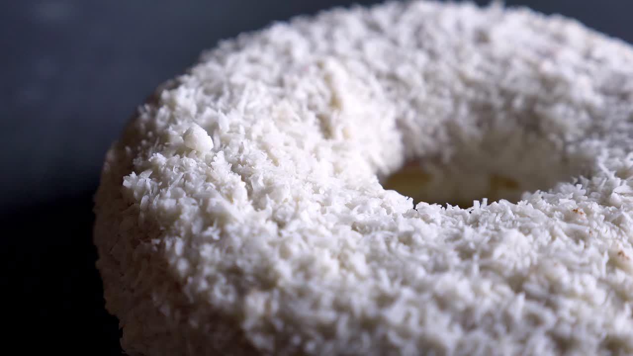 Close-up half-shot of tasty white donut with coconut chips spinning slowly on gray table background.