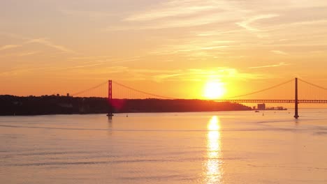 yellow ball of sun shines golden beam across pink orange water reflecting sky with backlit silhouette of suspension bridge