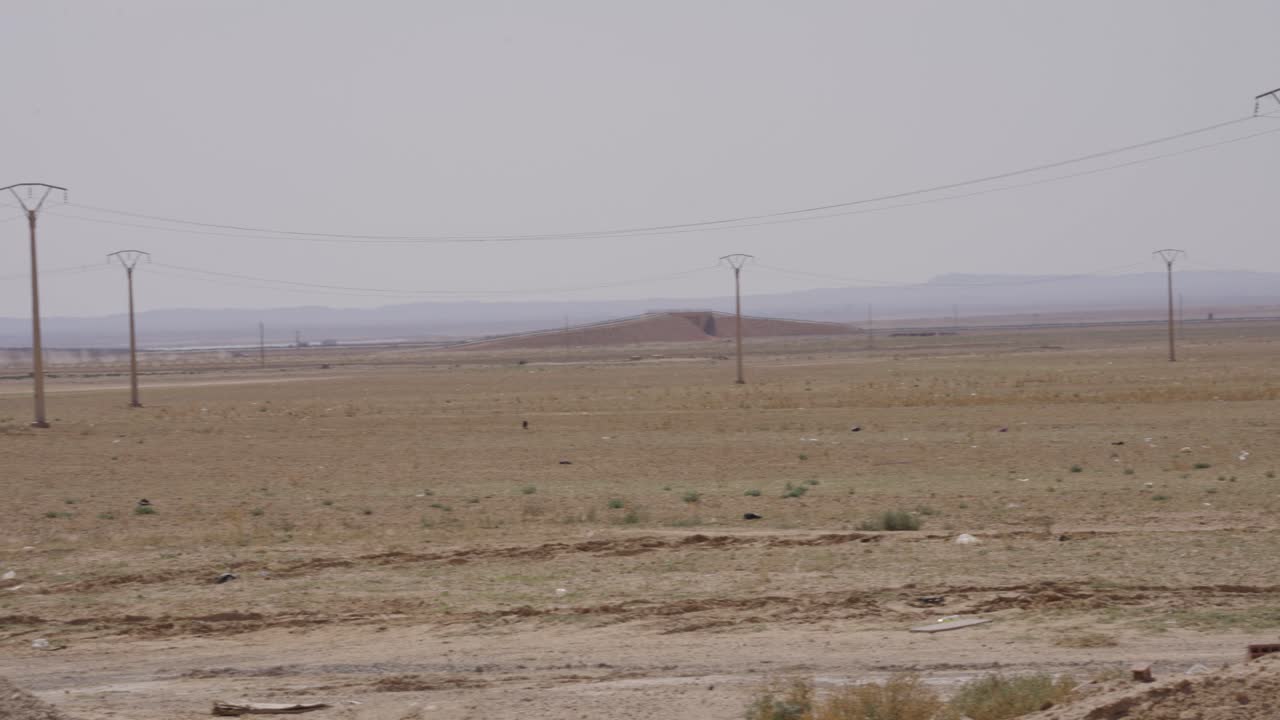 A wide shot of the hot, arid landscape of rural Algeria. The scene features a vast, dry plain, a remote village in the distance, and power lines under a hazy sky