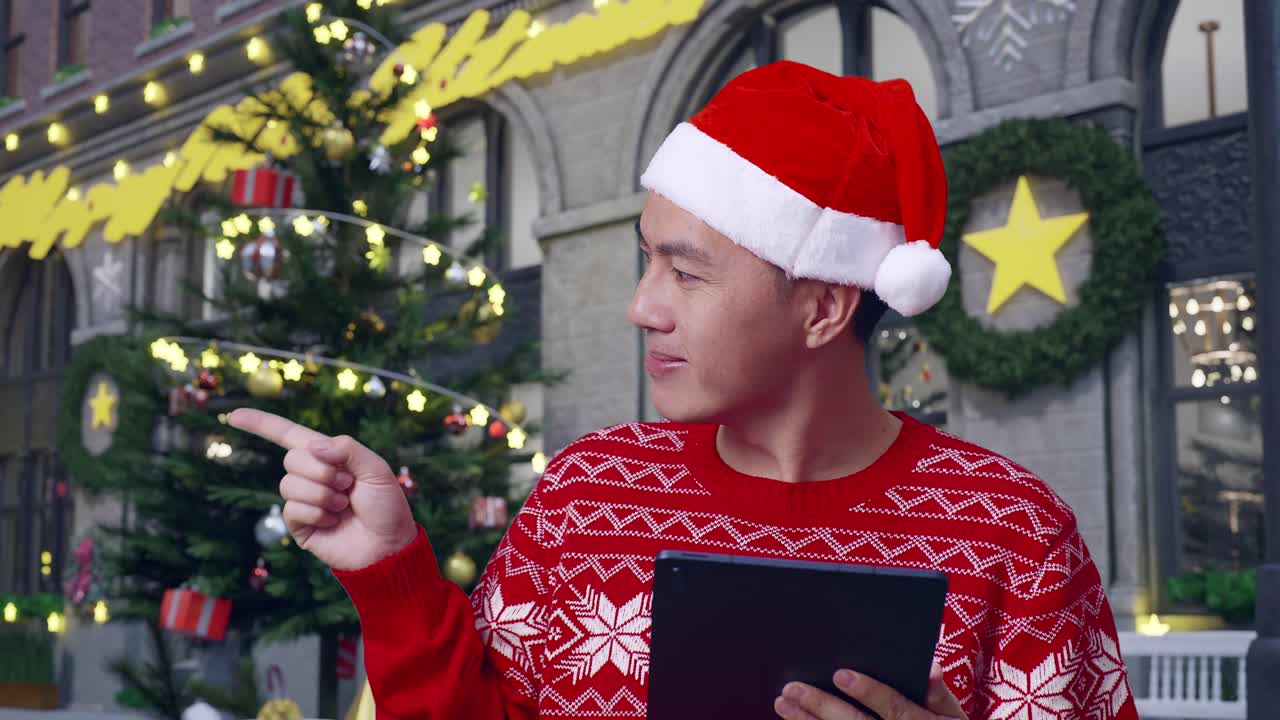 hombre con sombrero de papá noel sosteniendo la tableta frente a las decoraciones de navidad