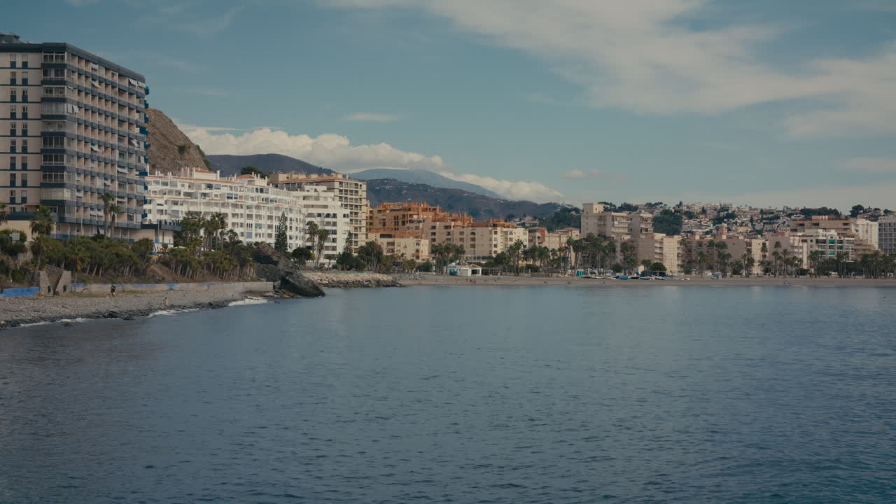 A closer view of Almuñécar’s beachfront with modern apartment buildings, palm trees, and distant hills under a soft blue sky
