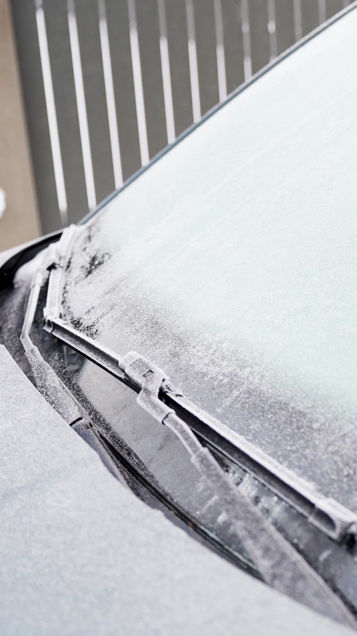 Close-up of a windshield wiper frozen under snow and frost on a parked car, showing icy winter conditions and the need for deicing before driving. Vertical b-roll.