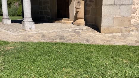 Filming in slow motion one of the entrances to a sanctuary we see the stone floor, the wooden porch supported by round granite columns, the wooden door, we see its garden area and a blue sky