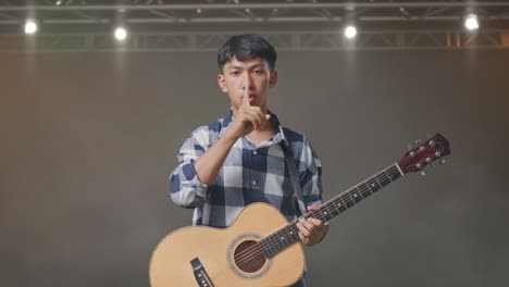 adolescente asiático con guitarra mostrando el gesto del silencio mientras está de pie en el escenario