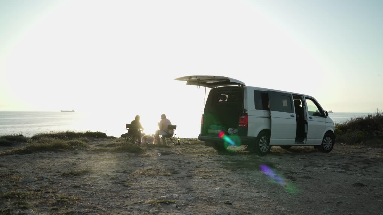 Two people sitting on the top of a hill in the sunset with their mini van car