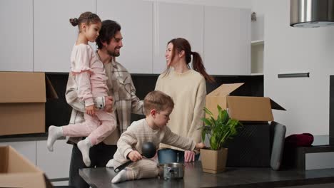 Happy-brunette-girl-in-a-beige-sweater-together-with-her-brunette-husband-in-a-plaid-shirt-who-is-holding-their-little-daughter-in-beige-clothes-talking-while-standing-in-the-kitchen-with-their-little-son-after-moving-to-a-new-apartment