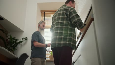 A-brunette-man-in-a-checkered-green-shirt,-together-with-his-elderly-boyfriend-with-gray-hair-and-a-lush-gray-beard-in-a-gray-T-shirt-are-cleaning-up-the-kitchen-after-preparing-lunch-together