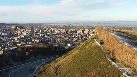 Toma-Aérea-De-Edimburgo-En-La-Nieve,-Con-El-Asiento-De-Arturo-Y-Los-Riscos-De-Salisbury-En-Primer-Plano,-Y-El-Castillo-De-Edimburgo-En-El-Fondo