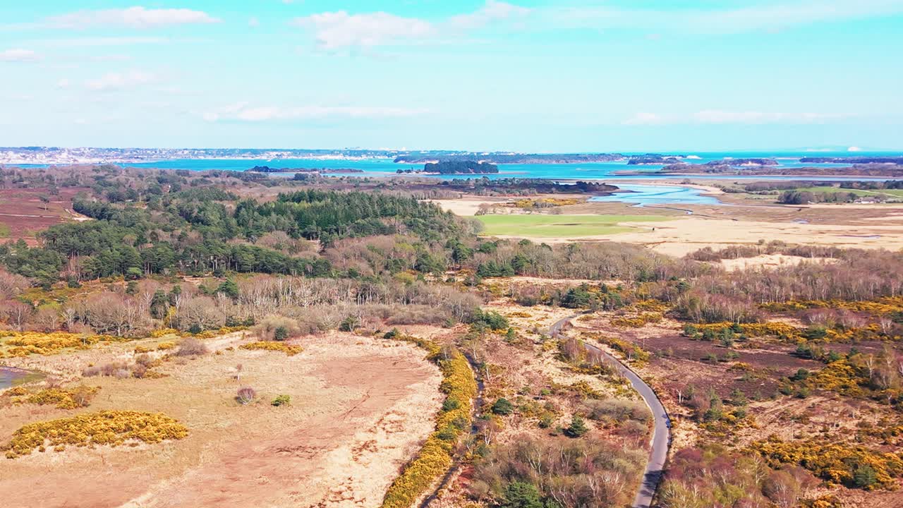 Luftfoto af hedeområde med snoet vej, der fører gennem landskabet mod en kystnær flodmunding og blåt vand under en klar himmel i Storbritannien.
