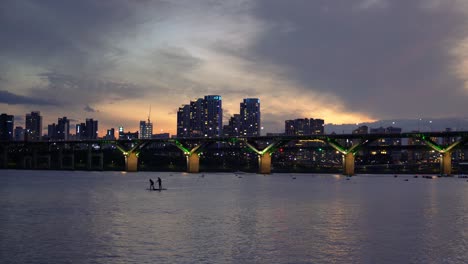 Silhouette-of-Two-People-Stand-Up-Paddleboarding-on-Han-River-at-Twilight,-Cheongdam-Bridge-with-Night-Cars-Traffic-and-Jamshil-District-of-Seoul-on-Colorful-Purple-Sky-Background,-Static-4k