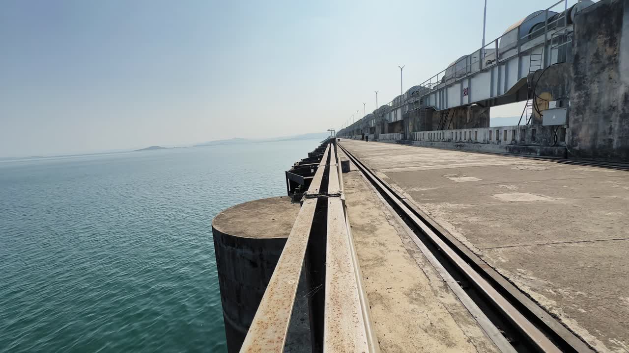 Kamera fährt von rechts nach links entlang eines indischen Dammwegs und enthüllt ruhiges blaues Stauseewasser, Betonstrukturen, Eisenbahnschienen und ferne Hügel unter klarem Tageslichthimmel