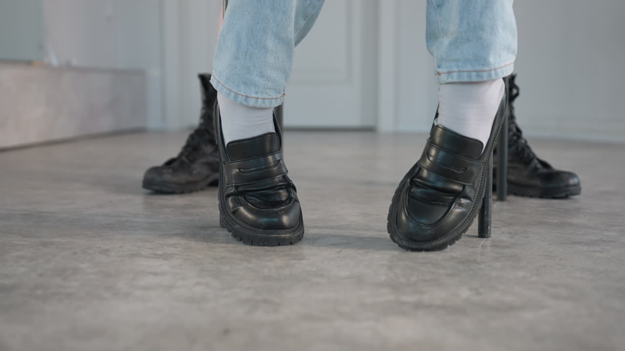 low angle closeup shows person seated in light denim raising polished black loafers above gray studio floor while another individual wearing rugged black combat boots strides past behind
