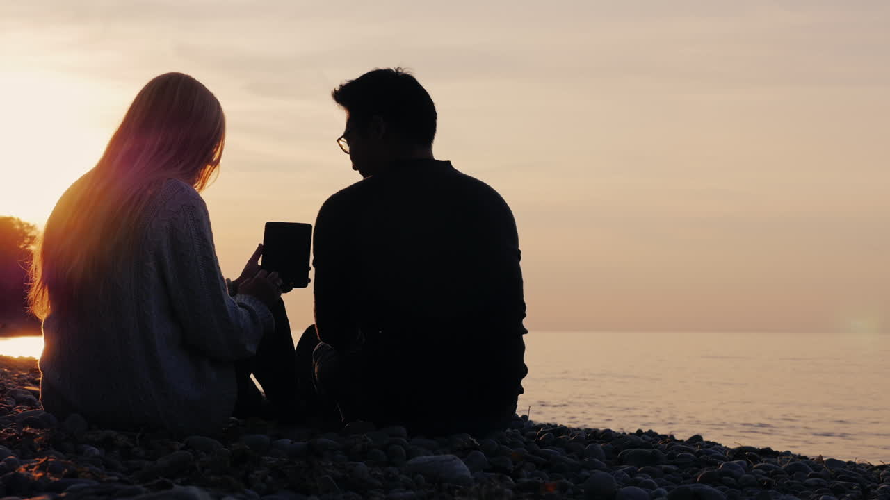 태블릿을 사용하여 호수 기슭에서 쉬고 있는 젊은 부부의 실루엣