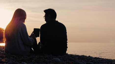 Silhouettes-of-a-young-couple-resting-on-the-shore-of-the-lake-using-a-tablet