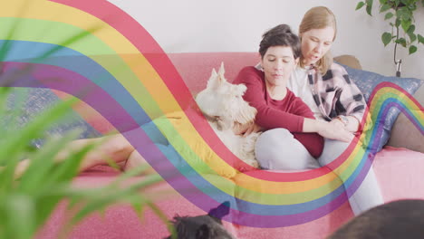 animation of pride rainbow band over happy caucasian lesbian couple relaxing on couch with pet dog