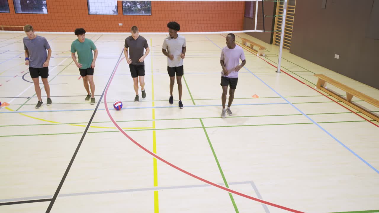 Diverses männliches Volleyballteam macht Ausfallschritte und geht zum Jogging bei Hütchen-Training mit Volleyball in der Turnhalle über.