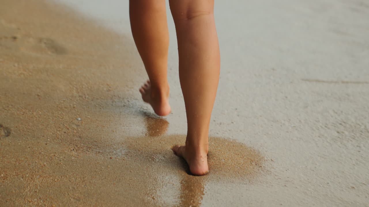Piedi che sguazzano nella sabbia bagnata, jogger giocoso che solleva schiuma e spruzzi, bagliore dell'ora d'oro, passo ritmico che lascia impronte fugaci, primo piano del movimento delle gambe che trasmette energia e un'estate spensierata