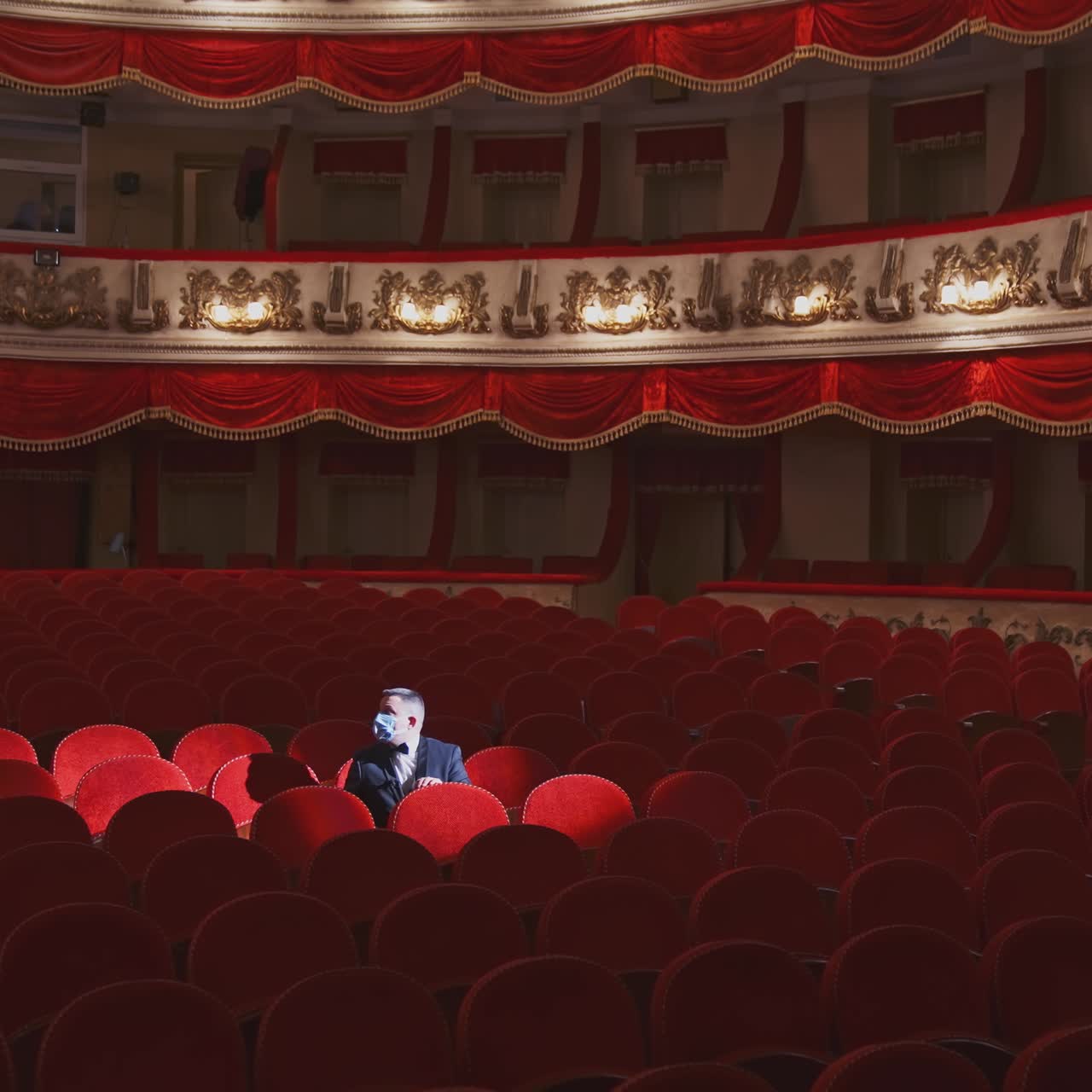 Rich man in mask in empty theater hall. Man spectator is sitting alone in large auditorium and looking to the sides. Consequences of pandemic.