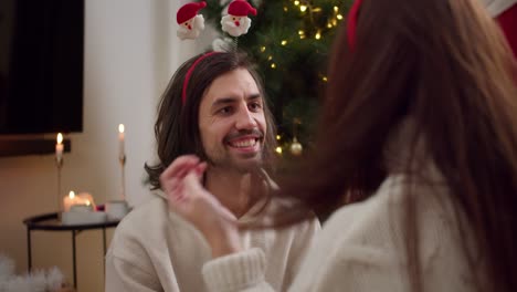 A-happy-brunette-guy-in-a-white-sweater-puts-a-New-Year's-hoop-on-the-head-of-his-brunette-girlfriend-in-a-white-sweater-near-a-green-Christmas-tree-decorated-with-a-yellow-garland-in-a-cozy-winter-home