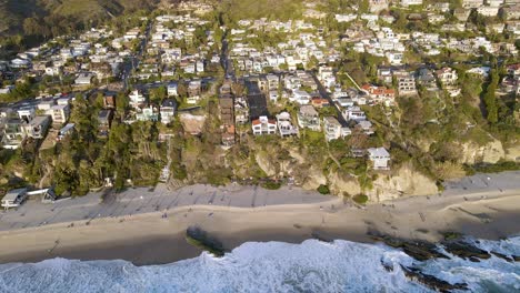 Aerial-top-down-shot-of-Thousand-Steps-Beach,-crashing-waves-with-foam-and-beautiful-cityscape-over-Laguna-City-during-sunset