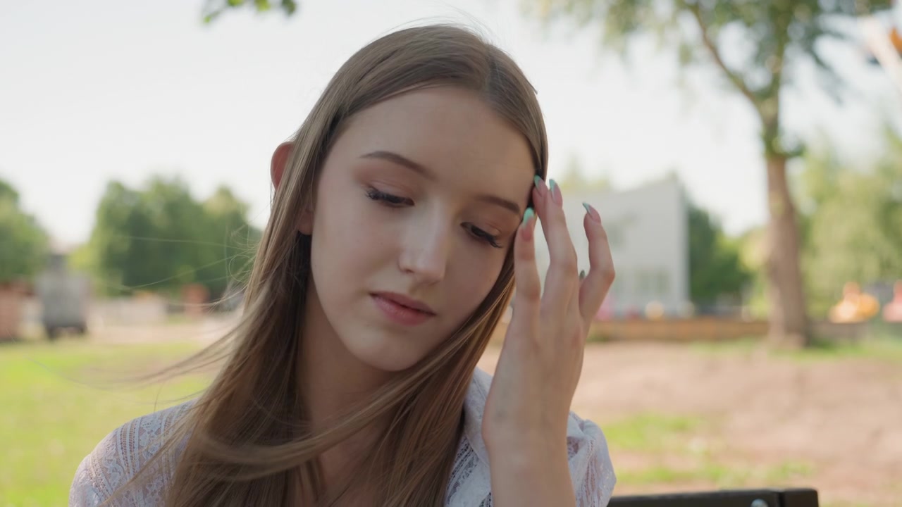 Primo piano di una giovane donna con i capelli lunghi seduta all'aperto in una luce diurna soffusa, che tiene palloncini colorati e aggiusta la sua posizione sulla panchina. La sua camicetta bianca presenta intricati dettagli in pizzo.