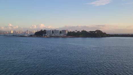 A beautiful scenic view across the tranquil ocean shows Naos Harbour Island on the Amador Causeway, with the modern Panama City skyline visible in the distance under a golden sky.