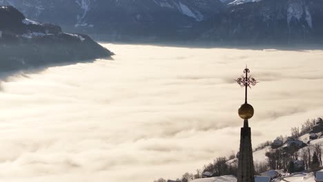 an aerial view of amden, switzerland, where a golden cross atop a church spire rises above a sea of fog, revealing a stunning panorama of the snow-covered valley below.