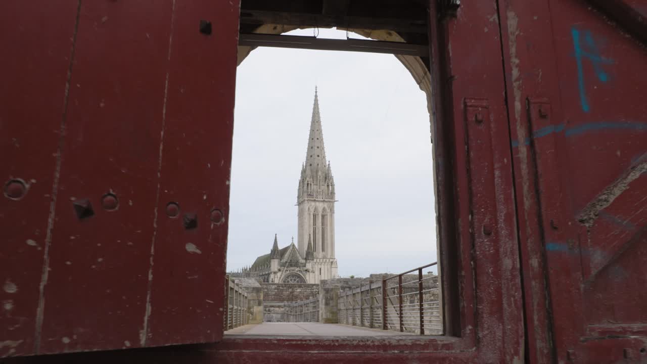 Low-angle moving footage capturing the majestic silhouette of the Saint-Pierre Cathedral in Caen, as seen from inside the Château de Caen. Framed by the ancient stone doorway of the castle