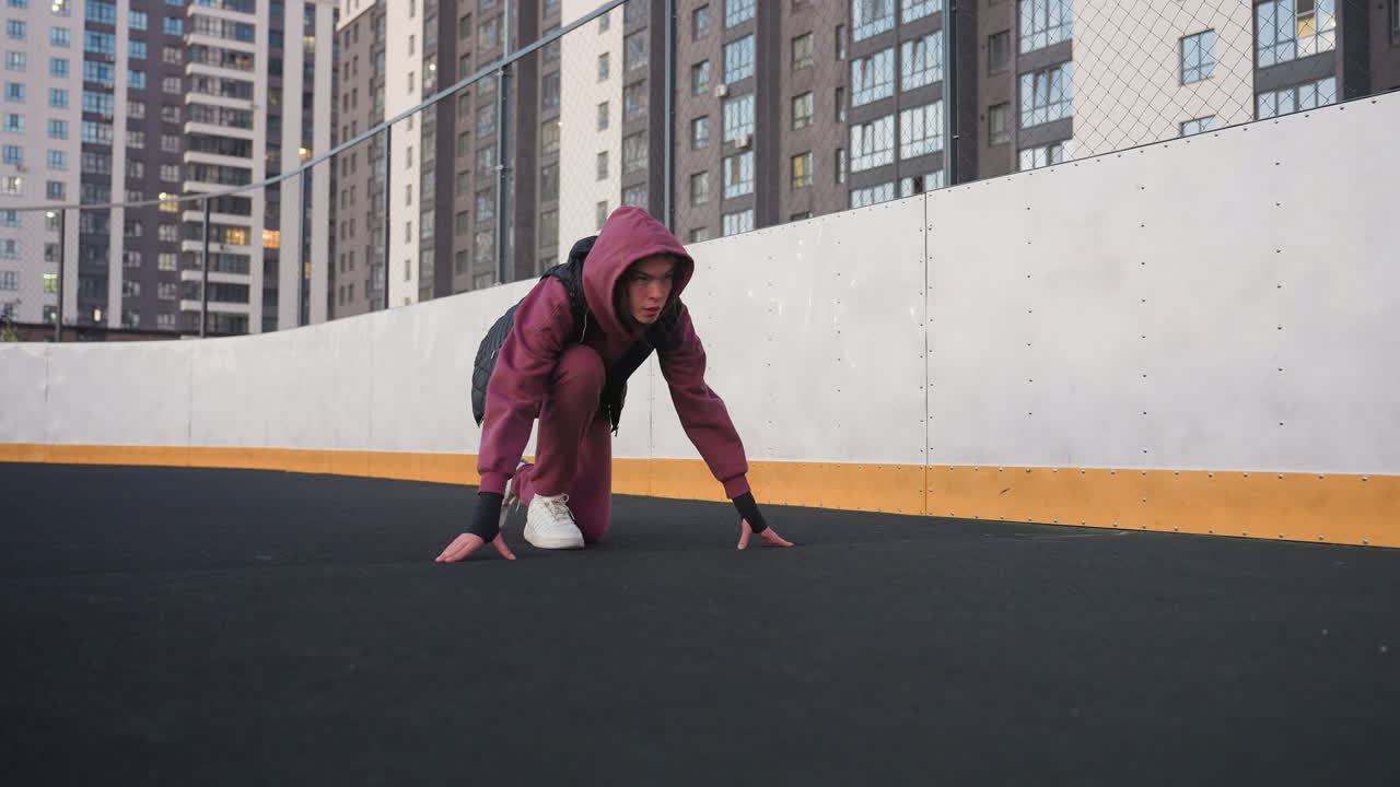 Gym goer kneeling in starting sprint position on black asphalt sports court next to white barrier topped with chain link fence in urban environment wearing maroon hoodie black vest and white sneakers