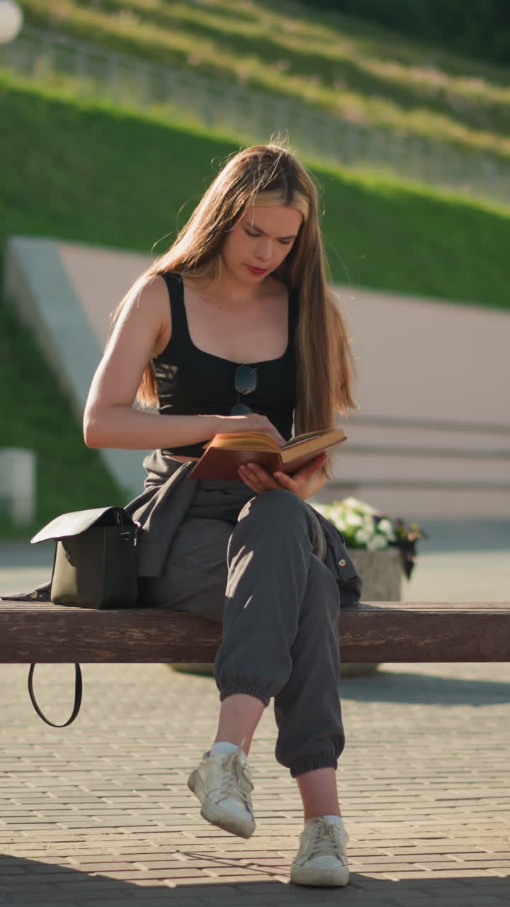 Lady seated outdoors with legs crossed flipping through a book, reading thoughtfully, the background features lamp poles and a blurred figure seated in the distance