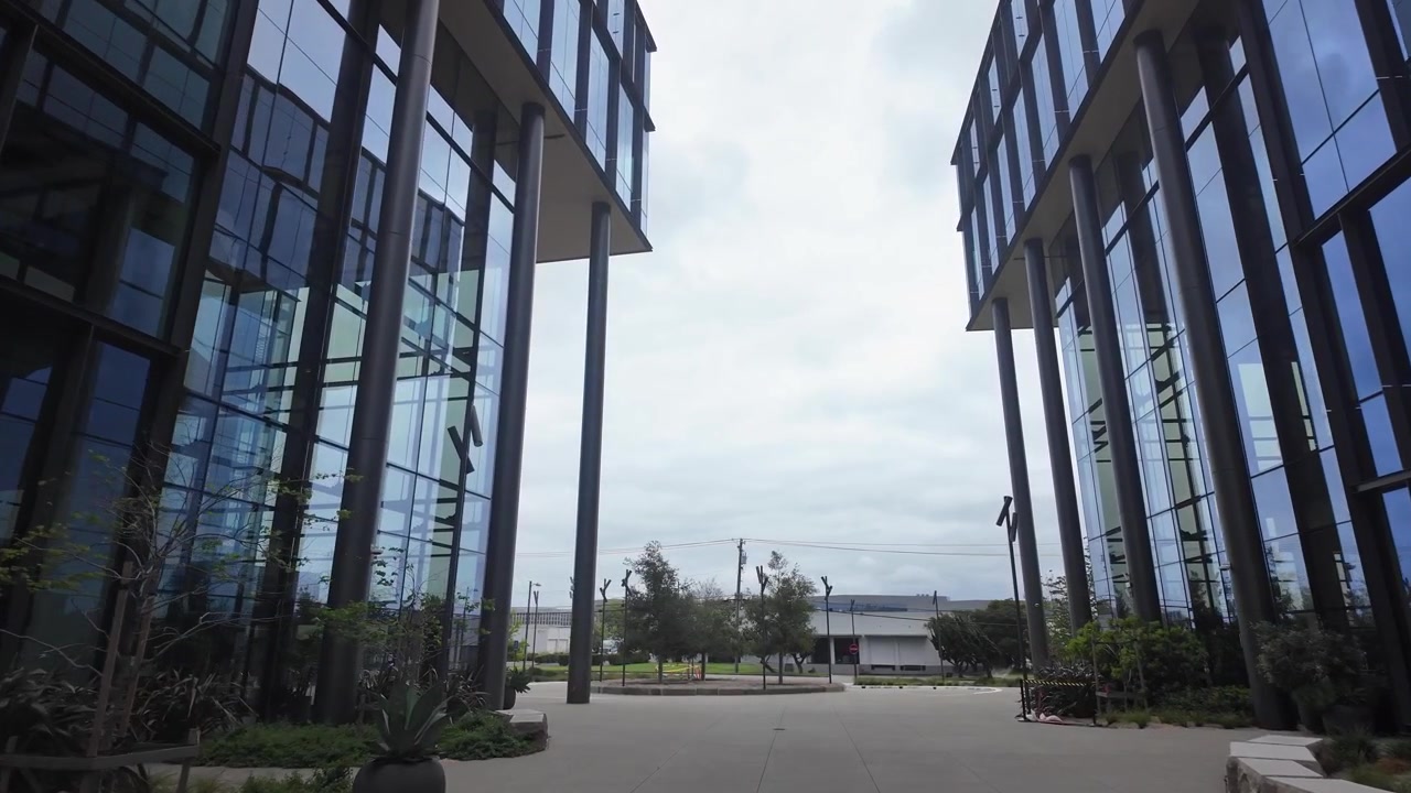 Movimiento lento hacia adelante a través de un patio vacío entre dos edificios de cristal contemporáneos, que forman parte de un campus de laboratorio médico en el sur de San Francisco, California, en un día nublado.