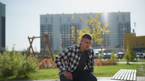 young man sits alone on wooden bench outdoors prepares to take pill under bright sunlight with trees swaying gently and cars parked in soft blur against backdrop of city apartment buildings