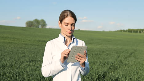 Caucasian researcher woman in white coat using tablet and looking at camera in the green field