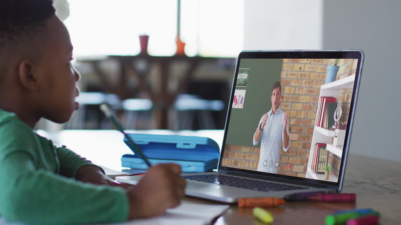 African american boy doing homework while having a video call with male teacher on laptop at home