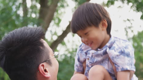 asian father lifting laughing son in  air as they play game in garden together