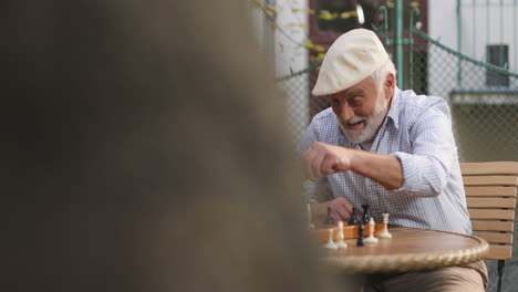 View-Over-The-Tree-On-The-Two-Male-Old-Cheerful-Friends-Sitting-On-A-Fresh-Air-In-The-Yard-And-Playing-Chess