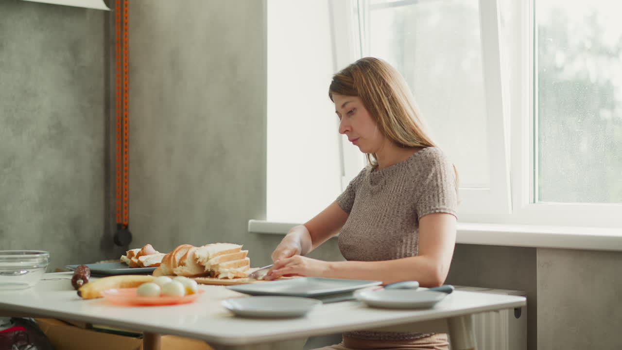 Medium shot of food preparer cutting sliced bread on plate with careful attention, table set with sausage banana eggs bowls knife nearby under bright window light creating calm culinary moment