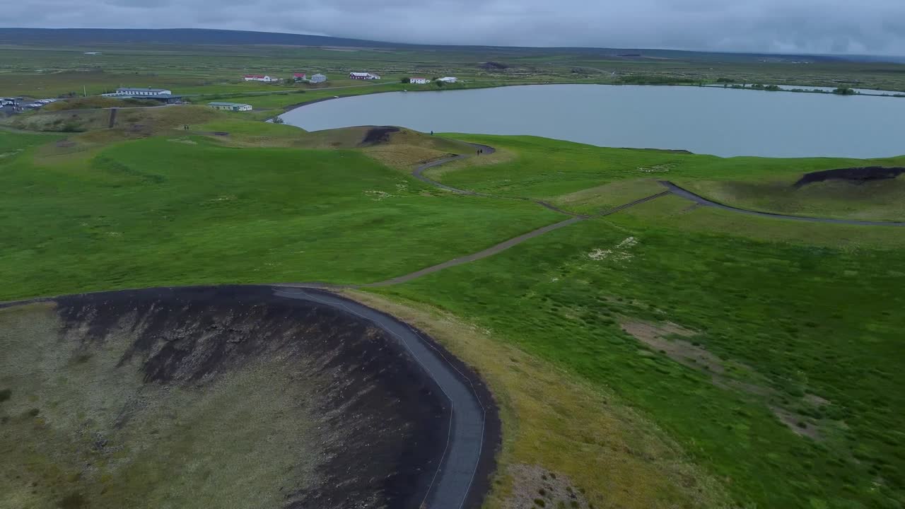 Ilmakuva jossa drone panoroi Myvatn-järven yllä. Järvessä näkyy vihreitä vulkaanisia pseudokraattereita ja tyyntä vettä. Laaja maisema esittelee Pohjois-Islannin ainutlaatuista geologiaa ja syrjäistä luonnonkauneutta.