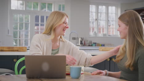 Teenage-daughter-giving-mother-gift-and-card-as-she-sits-at-kitchen-table-at-home-working-on-laptop---shot-in-slow-motion