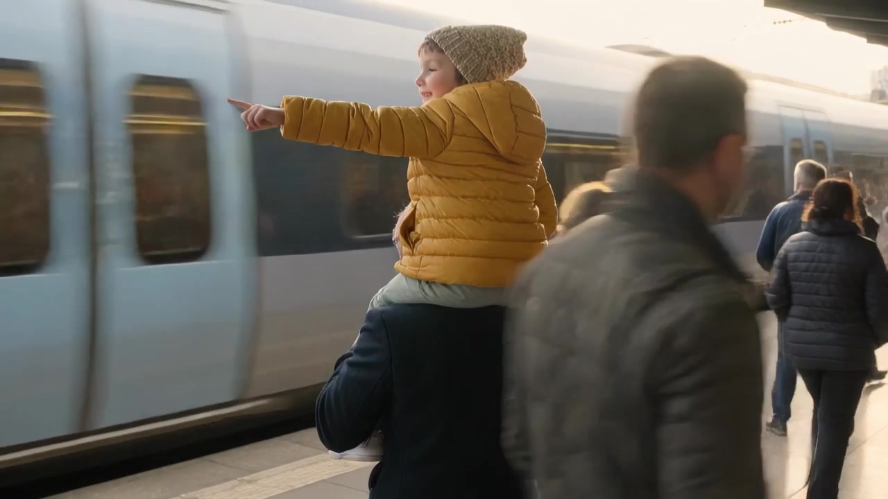 Walking man carrying boy in yellow puffer hat on platform holding legs, train passing, boy pointing