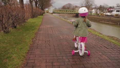 an adorable little girl in pink practicing how to ride a bicycle by the canal in spring after the rain - slow motion