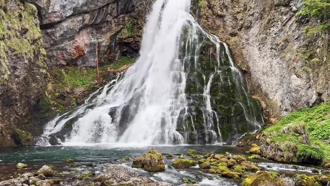 Vista dal basso della cascata di Golling dietro pietre chiare in primo piano, con una pozza d'acqua limpida, rocce muschiose e cascate a più livelli per riprese di viaggio panoramiche