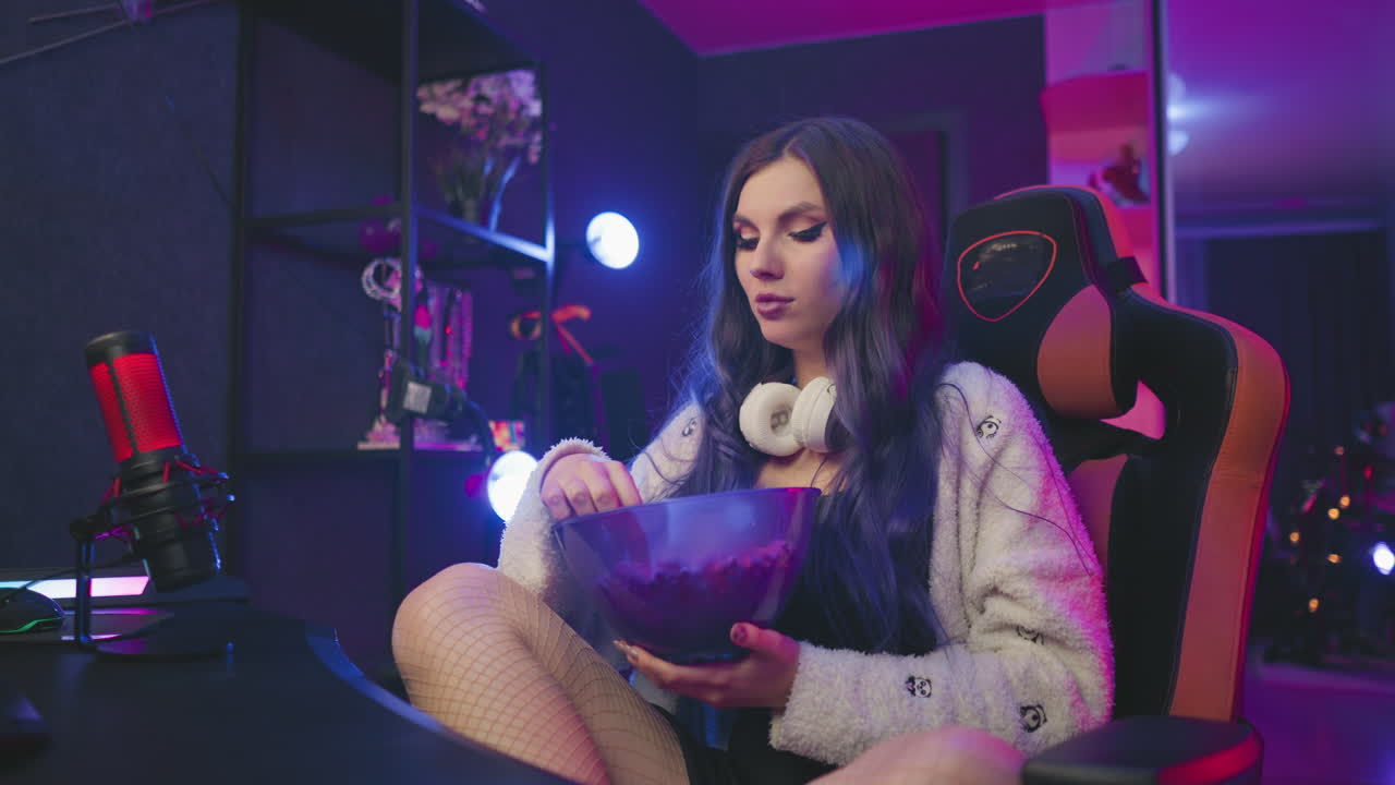 Woman with long hair sits comfortably in ergonomic gaming chair with legs crossed, eating popcorn from large bowl under soft lighting in modern studio filled with tech